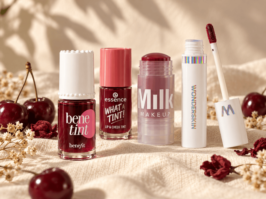 Benefit Benetint Dark Cherry next to essence What a Tint, Milk Makeup Cooling Water Jelly Tint and Wonderskin Wonder Blading Lip Stain on cream linen with fresh cherries