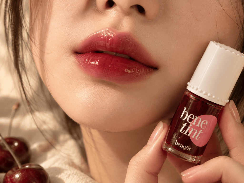 Close-up of soft, blurred black cherry lips with clear gloss, with a hand holding Benefit Benetint in Dark Cherry